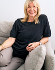 Woman sitting on a couch wearing a charcoal cashmere knit and gray pants, smiling.