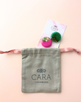 Two scarf brooches on a pink card with a gray 'Cara Cashmere' drawstring bag on a light pink background.
