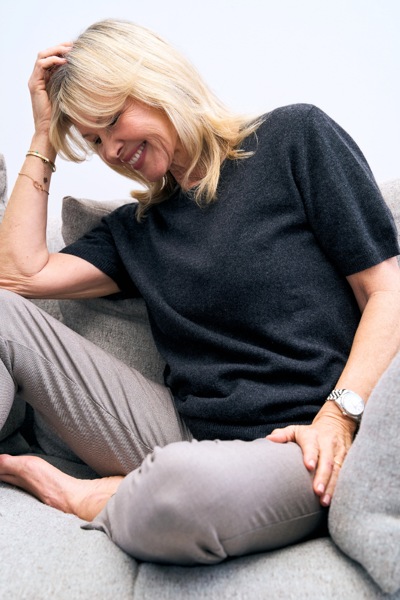 Woman sitting on a couch wearing a charcoal cashmere knit  and gray pants, smiling.