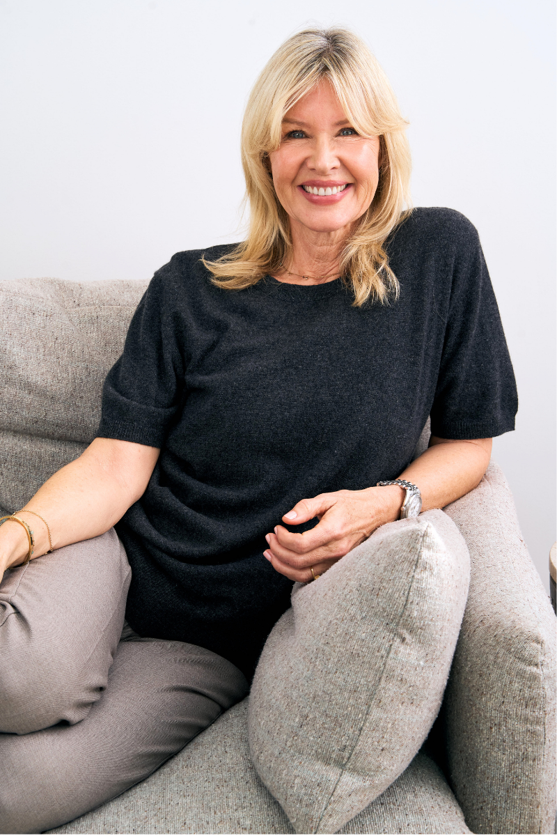 Woman sitting on a couch wearing a charcoal cashmere knit and gray pants, smiling.