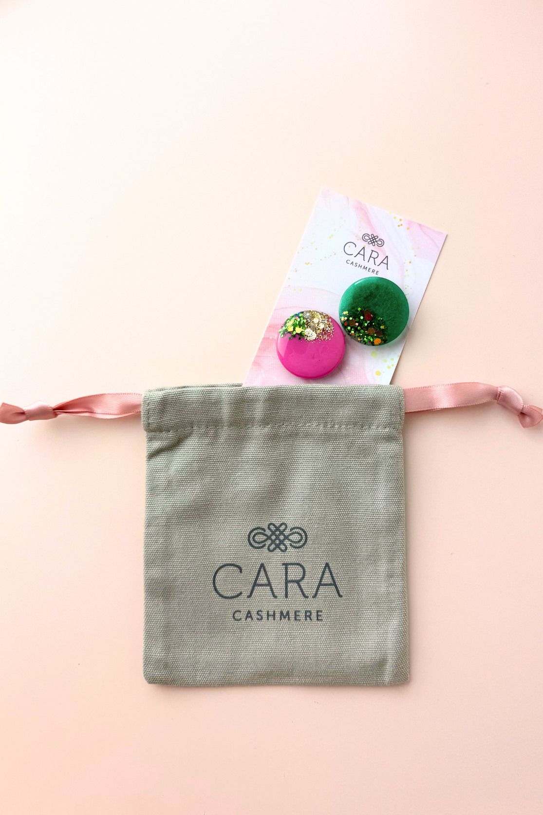 Two scarf brooches on a pink card with a gray 'Cara Cashmere' drawstring bag on a light pink background.